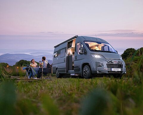 Ein Pärchen sitzt vor einem grauen Hobby Kastenwagen an einem Camping-Tisch in der Abenddämmerung.