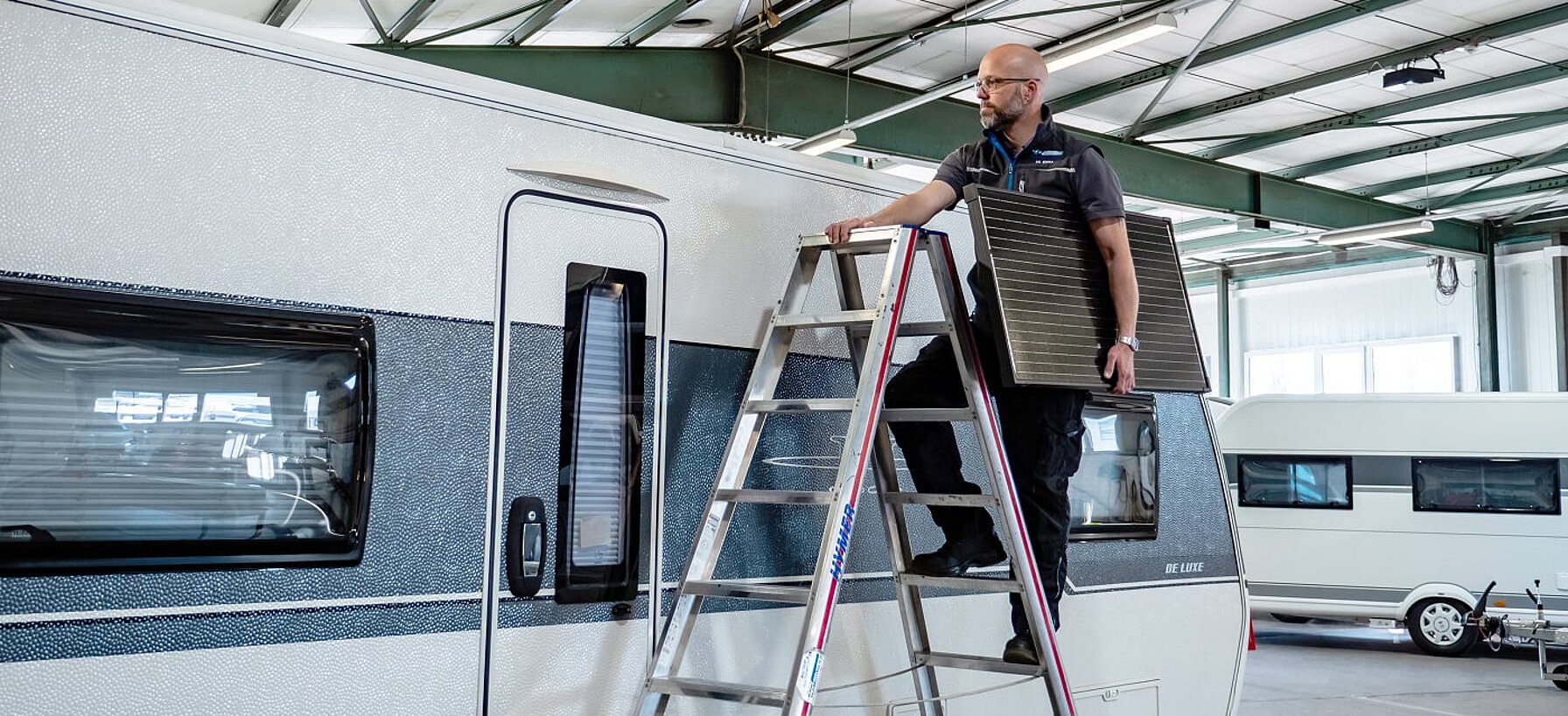 Werkstattmitarbeiter von Caravaning Center Schmidtmeier auf einer Leiter an einem Wohnwagen und einer Solar-Paneele unter dem Arm.