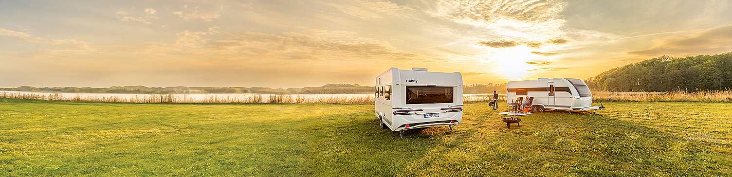 Zwei weiße Hobby Wohnwagen geparkt an einem See in einer sonnigen Umgebung. Dazwischen aufgebaute Camping-Garnitur und Grill mit verschiedenen Personen.