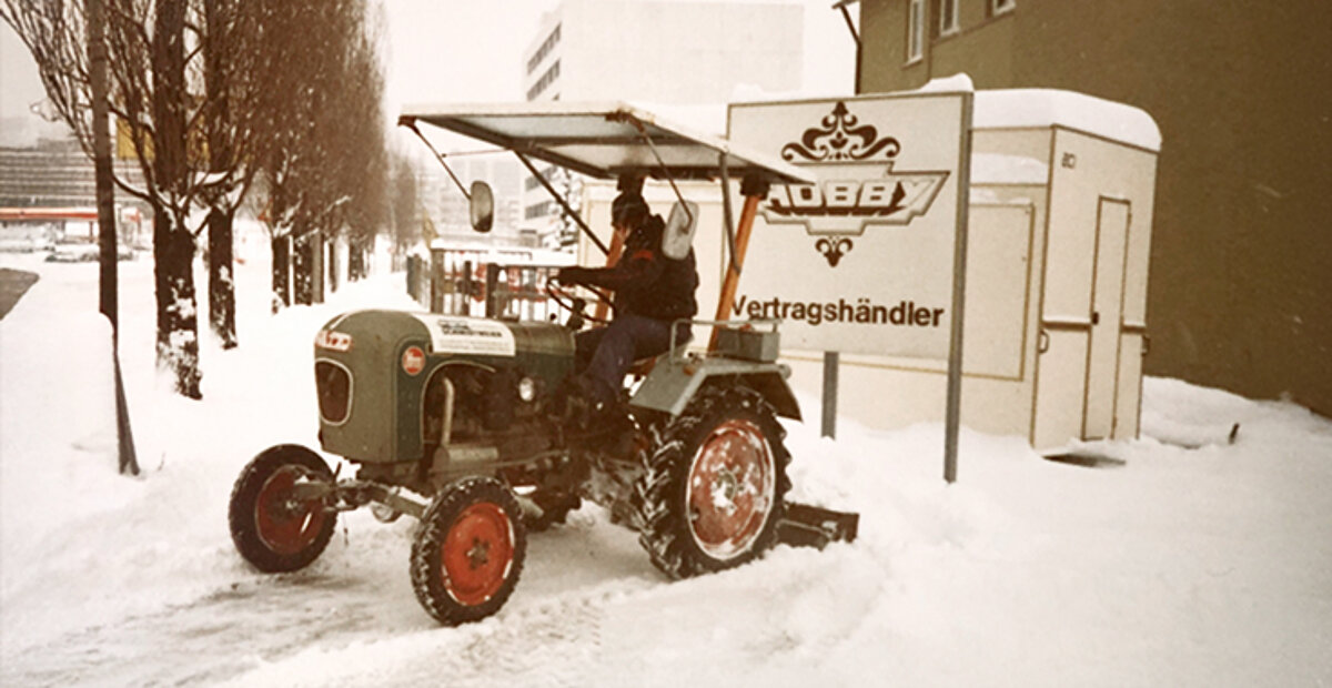 Sicht auf das Schild "Hobby Vertragshändler". Davor ein Trecker, der die schneebedeckte Außenfläche freiräumt.