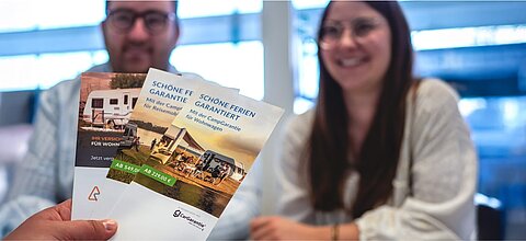 Ein glückliches Kunden-Pärchen im Hintergrund, davon eine Hand die drei Informationsbroschüren zum Thema Car-Garantie hält.