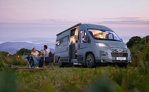 Dunkelgrauer Hobby Kastenwagen geparkt auf einem Feld in der Abenddämmerung. Davor ein Pärchen beim Abendessen am Campingtisch. ei
