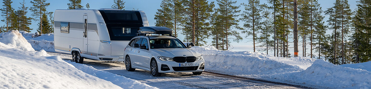 Ein weißer Wohnwagen gezogen von einem weißen PKW in einer Schneelandschaft.