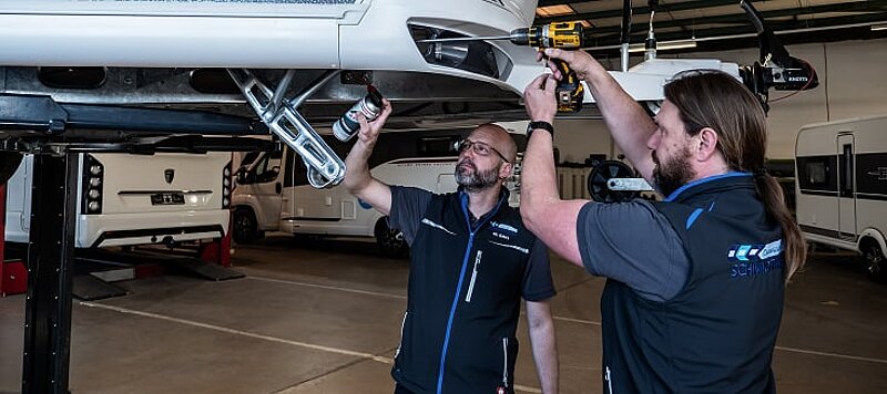 Zwei Werkstattmitarbeiter von Caravaning Center Schmidtmeier bei der Arbeit an einem Wohnwagen auf einer Hebebühne. Zwei Werkstattmitarbeiter von Caravaning Center Schmidtmeier bei der Arbeit an einem Wohnwagen auf einer Hebebühne.